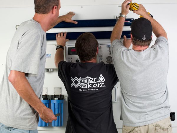 Three men from the WaterMakers team installing a Watermaker WMS-1000 system. One man is wearing a black t-shirt with the WaterMakers logo on it while holding the system in place. The man on the right is holding a drill.