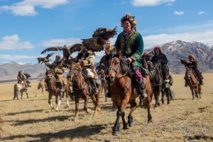 Mongolian eagle hunters riding to the festival on horses. There a white clouds in the sky and mountains in the distance.