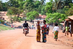 Two women standing in the street and talking in Gbarnga, Liberia. People are walking and riding motorized bikes in the distance